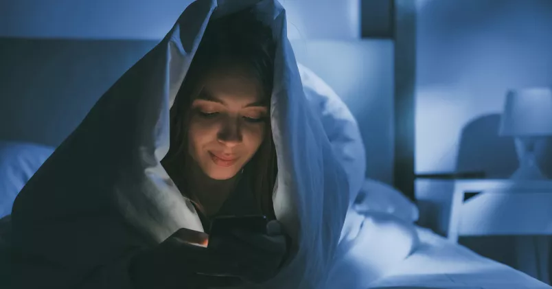 Kvinna ligger under täcket i sängen och kollar i sin mobil.