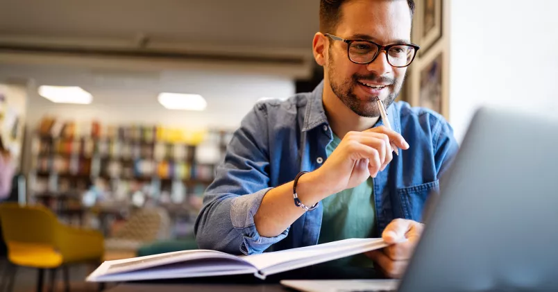 Man sitter i bibliotek med dator och bok.
