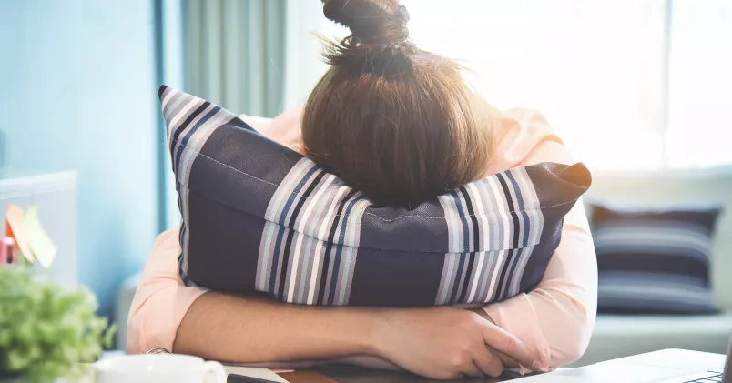 Kvinna vilar huvudet i kudde över sitt skrivbord.