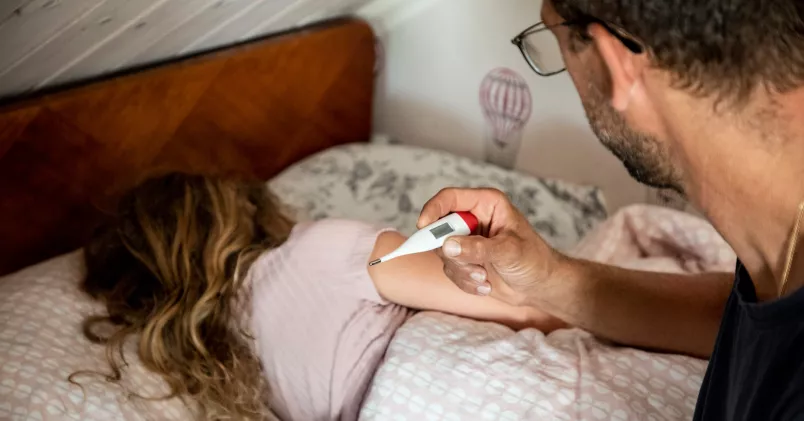Ett barn ligger i en säng. På sängkanten en pappa med en febertermometer i handen. 