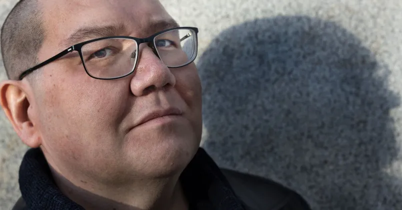 Arend Stührmann står mot en grå vägg och blickar in i kameran. Han bär svarta glasögon och svarta kläder, och ser allvarlig ut.