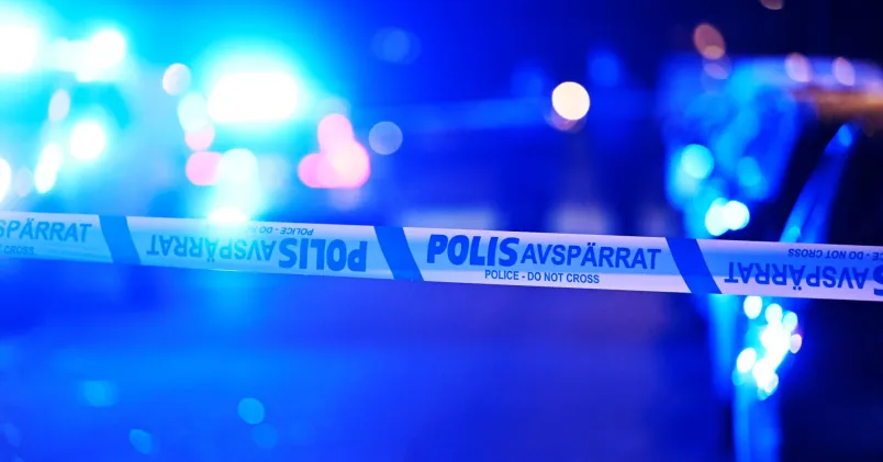 Avspärrningsband från polisen i förgrunden och blått utryckningsljus i bakgrunden – symbolbild för kriminalitet.