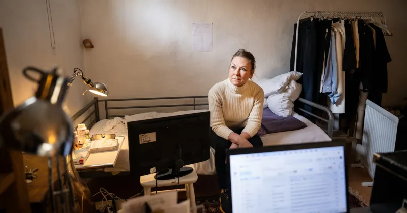 Maria Dahlin sitter i en säng i ett vitt källarrum. Framför henne står en uppfälld laptop. Hennes kläder hänger på en stång bredvid sängen.