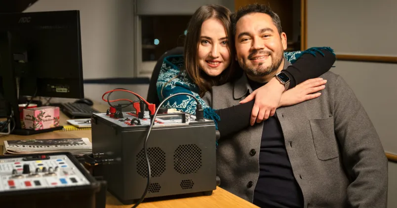 Kollegor som är ett par sitter tätt tillsammans på ett kontor, med armen om varandra bredvid mätutrustning – kärlek på jobbet.