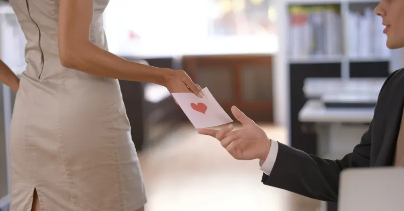 Person räcker över ett kuvert med hjärta till kollega på kontor – symbol för kärlek på jobbet.