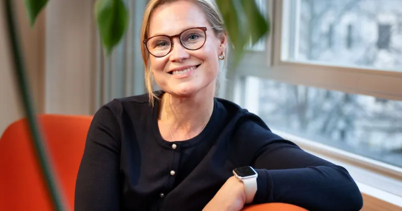 Blond kvinna med mörka glasögon och svarta kläder sitter i en orange soffa. Hon ser glad ut.