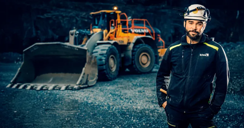 Marcus Linder jobbar som besiktningsingenjör. Står i Kiwa-märkta arbetskläder och skyddshjälm framför en hjullastare i en mörk bergtäkt.