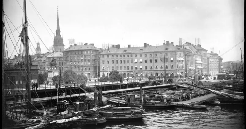 Småbåtar och bryggor i hamnen vid Slussen, med Stockholms bebyggelse och kyrktorn i bakgrunden.
