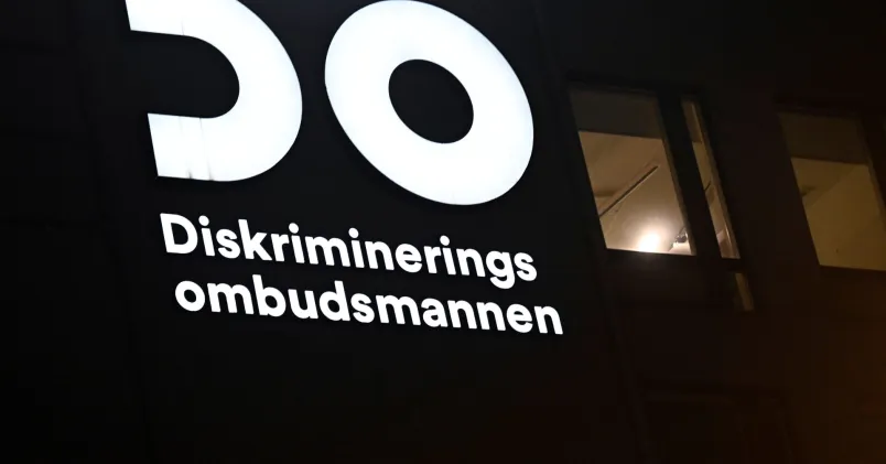 Upplyst skylt med DO:s logotyp och texten Diskrimineringsombudsmannen på en mörk husfasad.