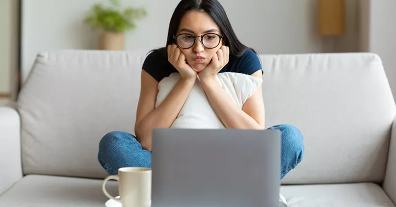 Kvinna sitter i soffan med huvudet i händerna framför laptop.