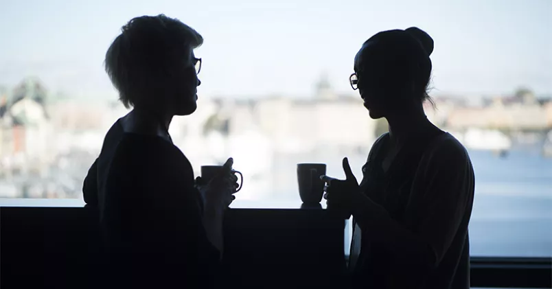 Två kvinnor står och pratar med kaffekoppar i händerna.