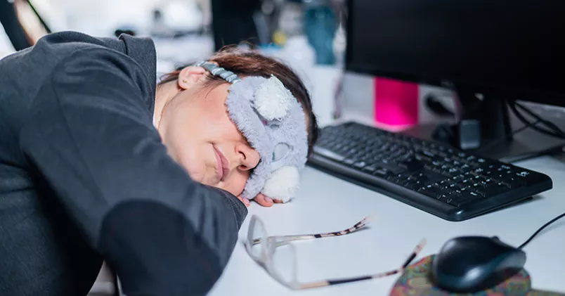 Kvinna iförd sovmask sover med huvudet på skrivbordet.