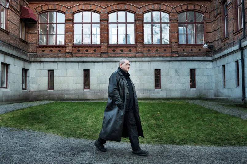 Arend Stührmann i lång svart läderrock promenerar ut ur bild. I bakgrunden syns en tegelvägg med stora fönster.