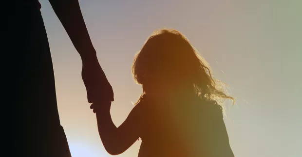 Siluett av barn som håller vuxen i hand i motljus.