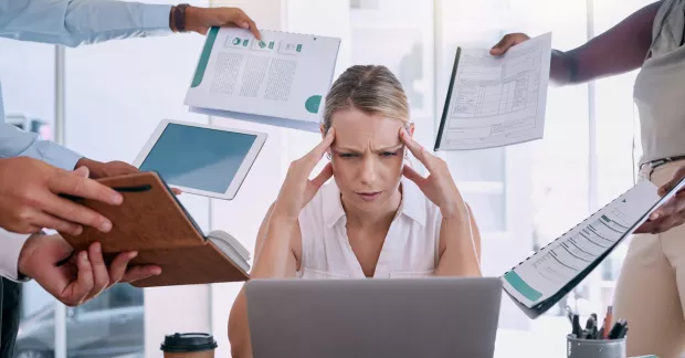 Kvinna vid bärbar dator omgiven av personer som alla ger henne olika papper. Håller sig för huvudet, stressar. 