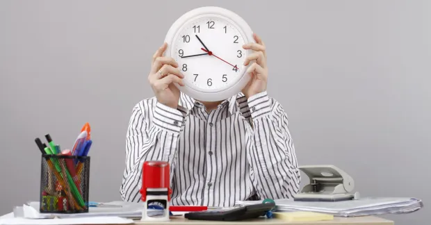 Arbetstidsbank - så funkar det. En man vid ett skrivbord med en stor klocka framför ansiktet. 