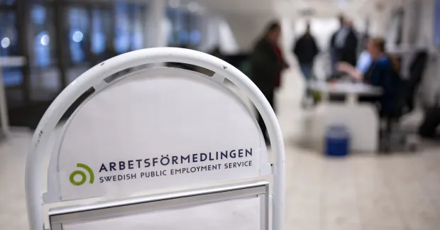 En skylt hos Arbetsförmedlingen, med jobbsökare i bakgrunden.