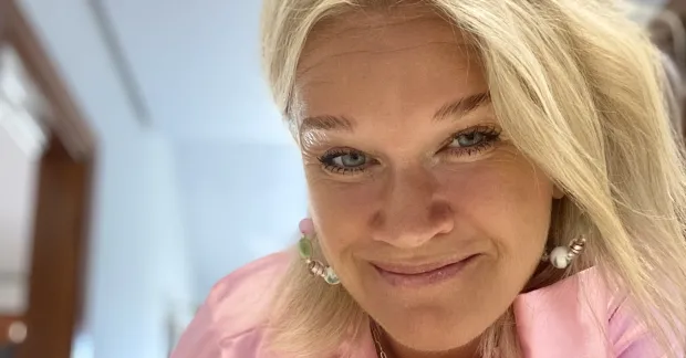 Tess Modig tittar in i kameran. Hon har långt blont hår och en rosa blus. Bakgrunden är lugn.