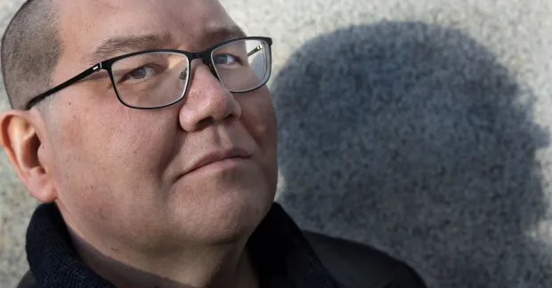 Arend Stührmann står mot en grå vägg och blickar in i kameran. Han bär svarta glasögon och svarta kläder, och ser allvarlig ut.