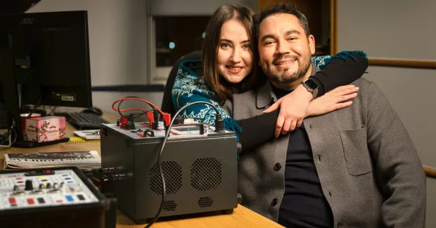 Kollegor som är ett par sitter tätt tillsammans på ett kontor, med armen om varandra bredvid mätutrustning – kärlek på jobbet.