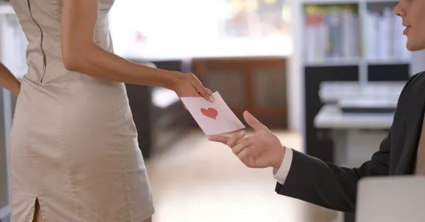 Person räcker över ett kuvert med hjärta till kollega på kontor – symbol för kärlek på jobbet.