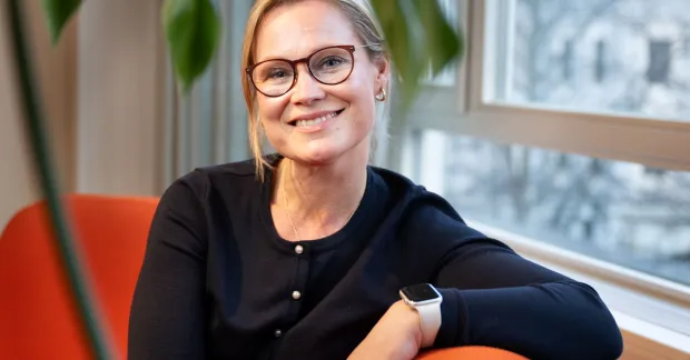 Blond kvinna med mörka glasögon och svarta kläder sitter i en orange soffa. Hon ser glad ut.