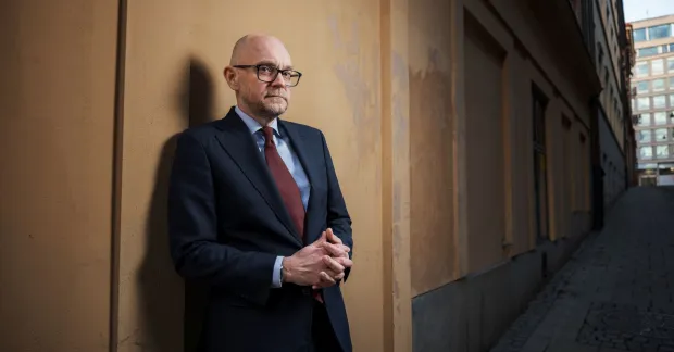 Johan Britz står lutad mot en gul stenvägg. Han bär glasögon och mörk kostym