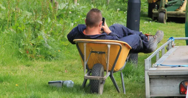 Lat på jobbet. En person lutar sig tillbaka i en skottkärra utomhus och håller en mobiltelefon.