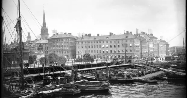 Småbåtar och bryggor i hamnen vid Slussen, med Stockholms bebyggelse och kyrktorn i bakgrunden.