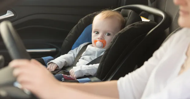 Litet barn fastspänt i ett babyskydd i baksätet på en bil medan en vuxen person sitter vid ratten.