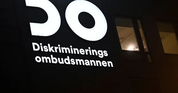 Upplyst skylt med DO:s logotyp och texten Diskrimineringsombudsmannen på en mörk husfasad.