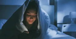 Kvinna ligger under täcket i sängen och kollar i sin mobil.