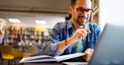Man sitter i bibliotek med dator och bok.