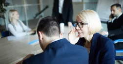 Kvinna i övre medelåldern viskar till manlig kollega vid konferensbord.