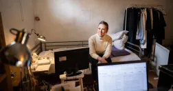 Maria Dahlin sitter i en säng i ett vitt källarrum. Framför henne står en uppfälld laptop. Hennes kläder hänger på en stång bredvid sängen.