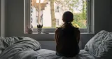 En person sedd bakifrån sitter på en säng och tittar ut genom ett fönster i ett sovrum. Bilden används som illustration till artikel om hemmasittare och press i arbetslivet.
