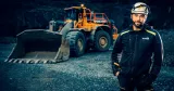 Marcus Linder jobbar som besiktningsingenjör. Står i Kiwa-märkta arbetskläder och skyddshjälm framför en hjullastare i en mörk bergtäkt.