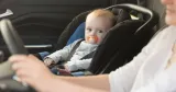 Litet barn fastspänt i ett babyskydd i baksätet på en bil medan en vuxen person sitter vid ratten.